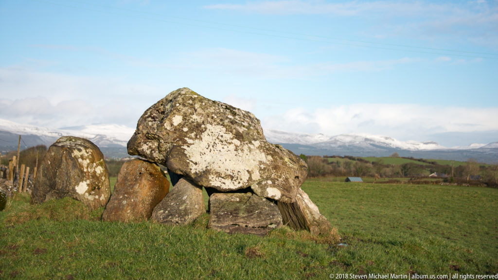 Oblique view of a large lichen-covered capstone balanced on upright stones in open pasture.