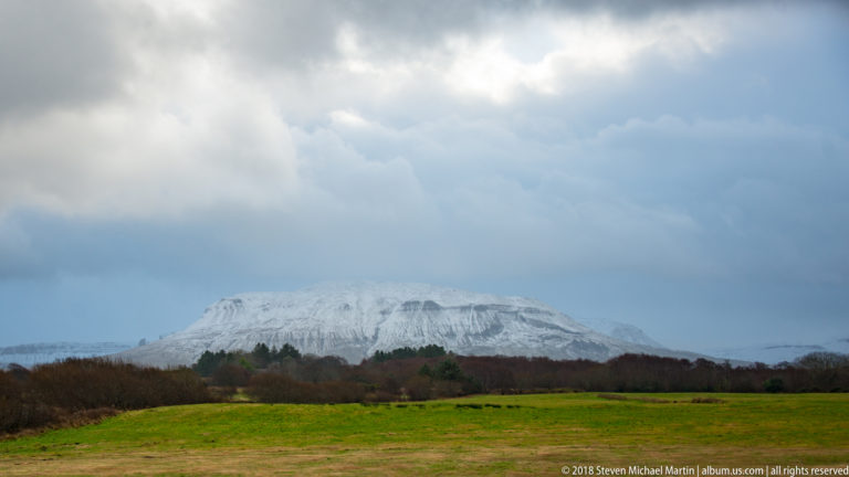Snow-covered flat-topped mountain beyond bright green pasture and dark winter vegetation beneath broken clouds.
