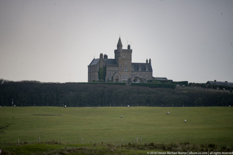 Distant castle-like historic building with a tall central tower beyond hedges and a sheep-dotted pasture.