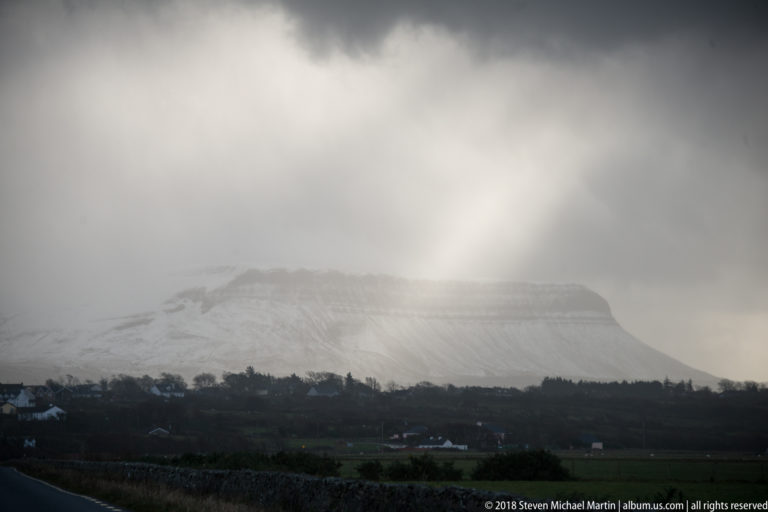 Snow-covered flat-topped mountain under dark cloud and mist, with bright light breaking over the summit.