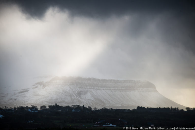 Snow-covered flat-topped mountain partly veiled by cloud, with a pale shaft of light crossing the upper slopes.