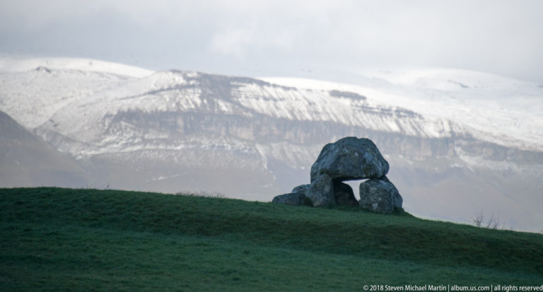 Small capstone tomb on a grassy ridge with a snow-covered mountain rising directly behind.