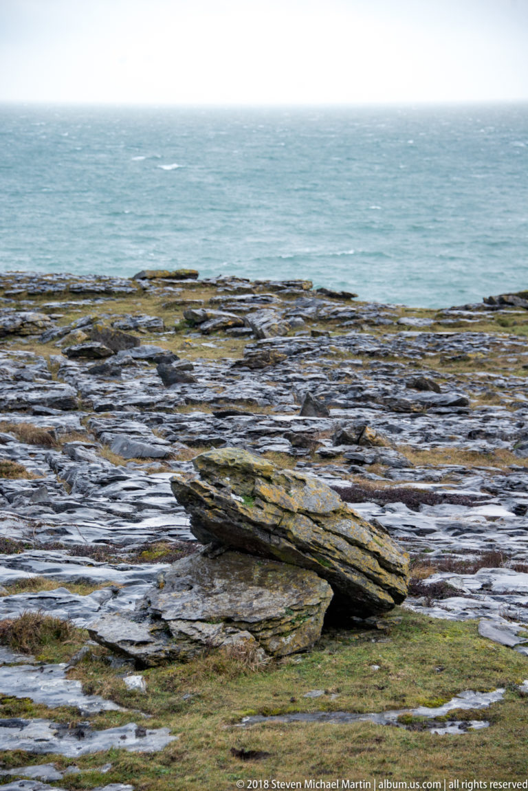 Long tilted limestone slab rising from flat coastal pavement, with sparse grass and the Atlantic on the horizon.