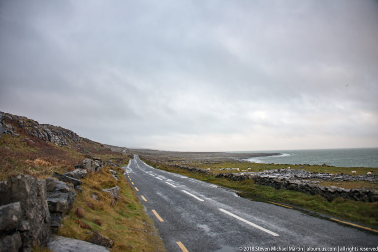 Wet two-lane road running between rocky slopes and low stone walls toward a distant Atlantic shoreline under gray cloud.
