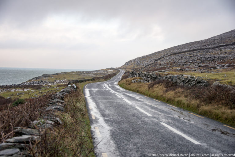 Wet narrow road winding between dry-stone walls and a bare limestone hillside, with the Atlantic visible to the left.