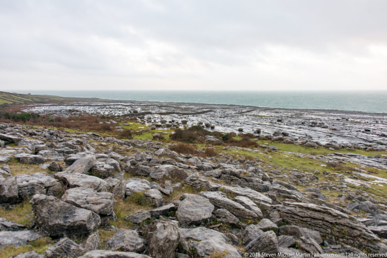 Broad fractured limestone pavement with scattered boulders and sparse vegetation stretching toward a blue-gray Atlantic horizon.