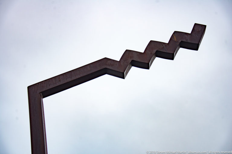 Dark angular metal sculpture with a vertical support and stepped zigzag upper section against a pale gray sky.