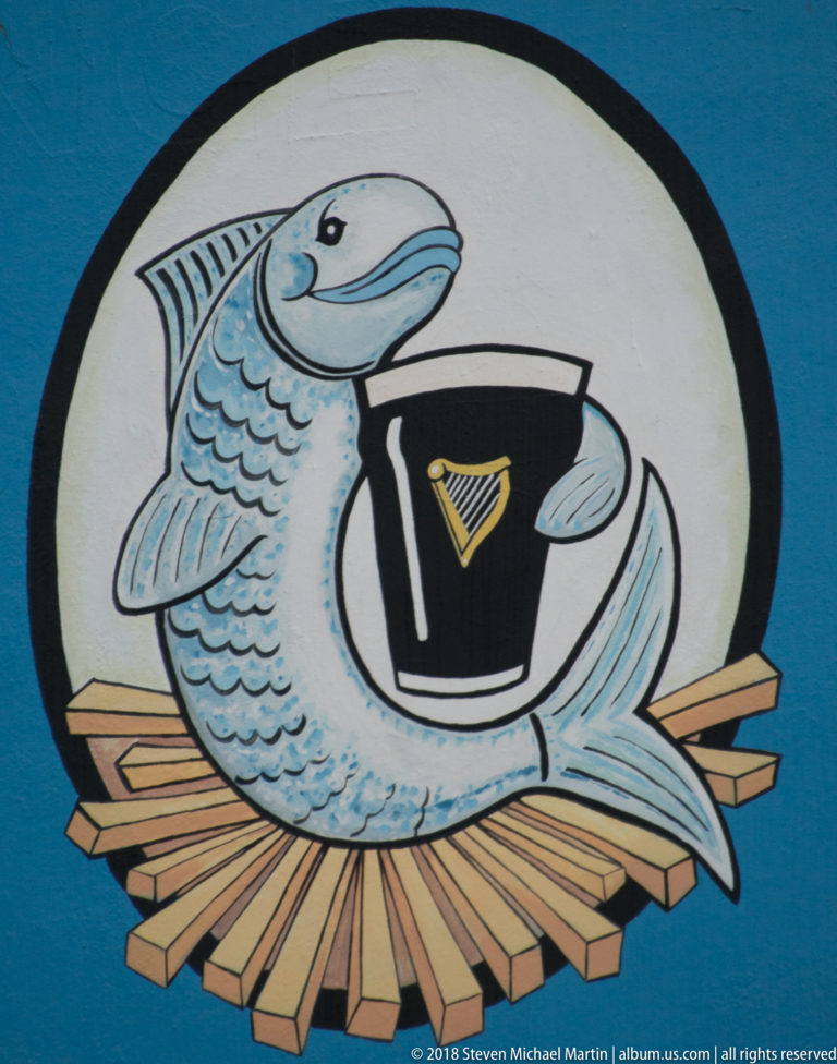 Stylized fish curled around a black pint glass with a gold harp, painted inside a white circle on a blue wall.