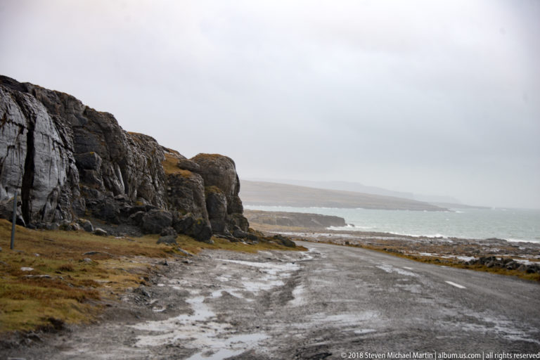 Wet coastal lane running beside a dark rock face, with low Atlantic surf and misty headlands to the right.