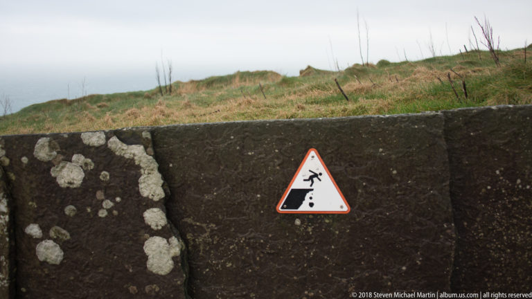 Red-bordered triangular warning sign of a person falling from a cliff, fixed to a lichen-covered concrete wall.