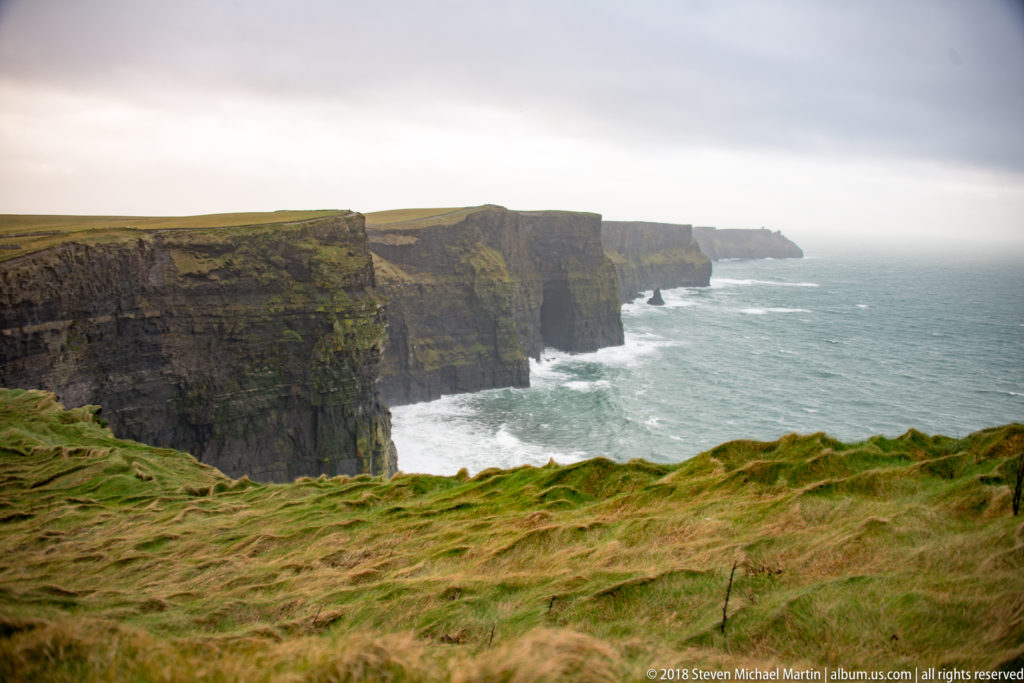 Layered dark Cliffs of Moher receding into mist above white Atlantic surf, with windblown green grass in the foreground.