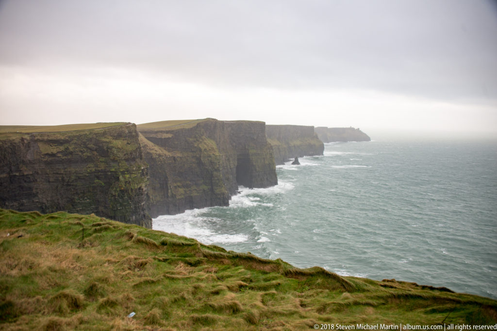 Grass-topped Cliffs of Moher extending into mist beside a wide expanse of gray Atlantic water and white surf.