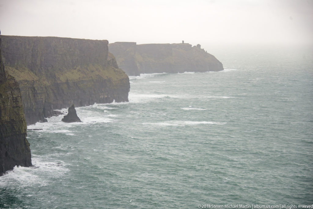 Narrow sea stack below the dark Cliffs of Moher, with foaming Atlantic surf and misty headlands beyond.
