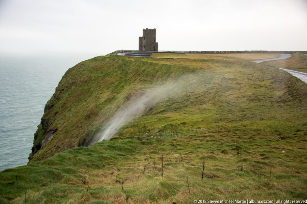 O'Brien's Tower on a green cliff-top promontory above the Atlantic, with windblown spray or rain crossing the slope.