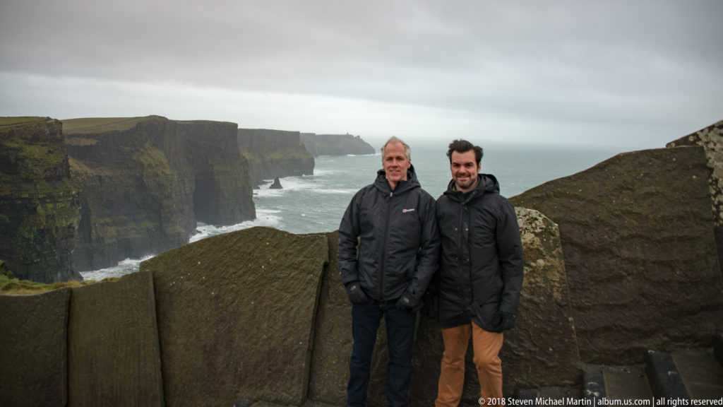 Two men in dark jackets posing at a stone viewpoint barrier, with the Cliffs of Moher and Atlantic surf behind them.
