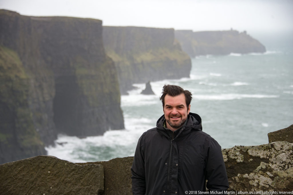 Smiling man in a black jacket at a stone viewpoint, with the Cliffs of Moher and white Atlantic surf behind him.