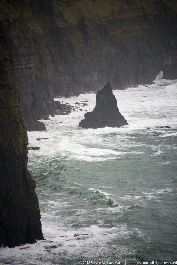 Dark sea stack surrounded by white Atlantic surf below steep, layered coastal cliffs at the Cliffs of Moher.