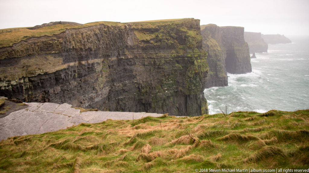Grass-topped Cliffs of Moher recede in a long sequence of sheer rock faces beside a misty Atlantic Ocean.