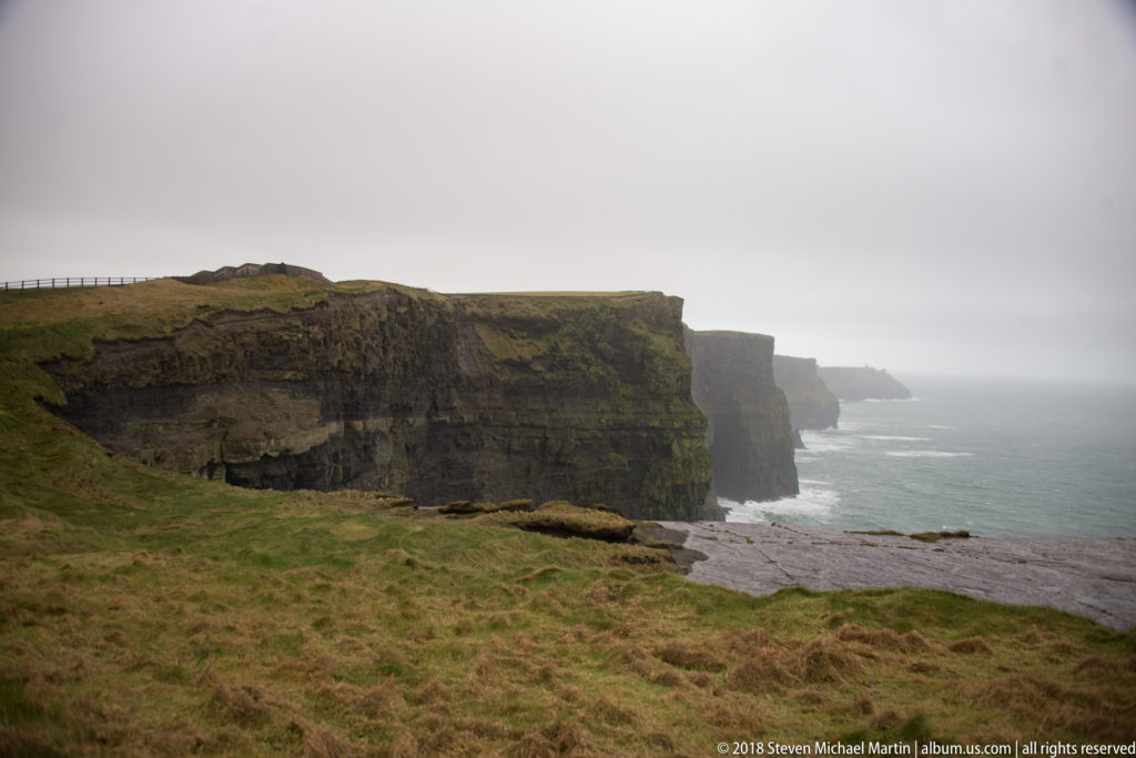 Broad grassy foreground leads to layered sea cliffs receding into gray mist along the Cliffs of Moher coast.
