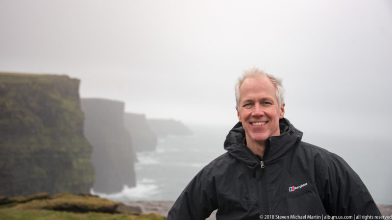 Smiling gray-haired visitor in a black jacket stands before misty Cliffs of Moher and Atlantic surf.