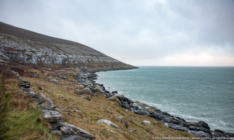Sloping rocky coast with brown grass and scattered boulders descending to blue-green Atlantic water under heavy cloud.