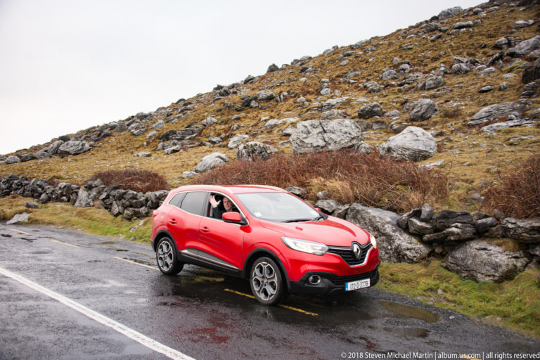 Red Renault crossover on a wet rural road beside a dry-stone wall and boulder-strewn hillside.