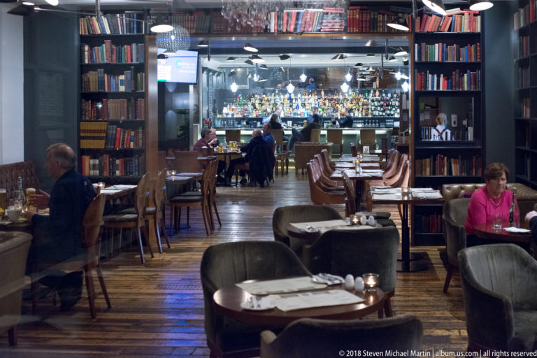 Hotel restaurant interior with book-lined walls, set tables, upholstered chairs, and illuminated bar beyond.