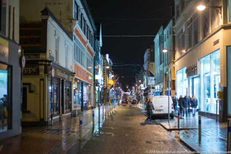 Wet nighttime commercial street with lit shops, pedestrians, traffic lights, and a white van.