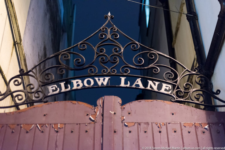 Decorative wrought-iron ELBOW LANE arch with scrollwork spanning a narrow passage.