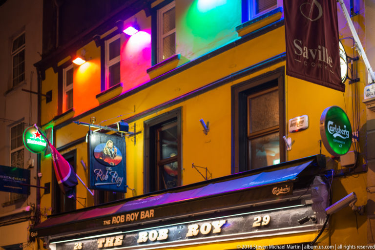 Bright yellow Rob Roy Bar façade with multicolored lighting, beer signs, and illuminated pub name.