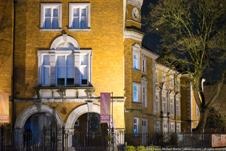 Yellow-brick historic building with arched entrance, sash windows, stone trim, iron fence, and tree at night.