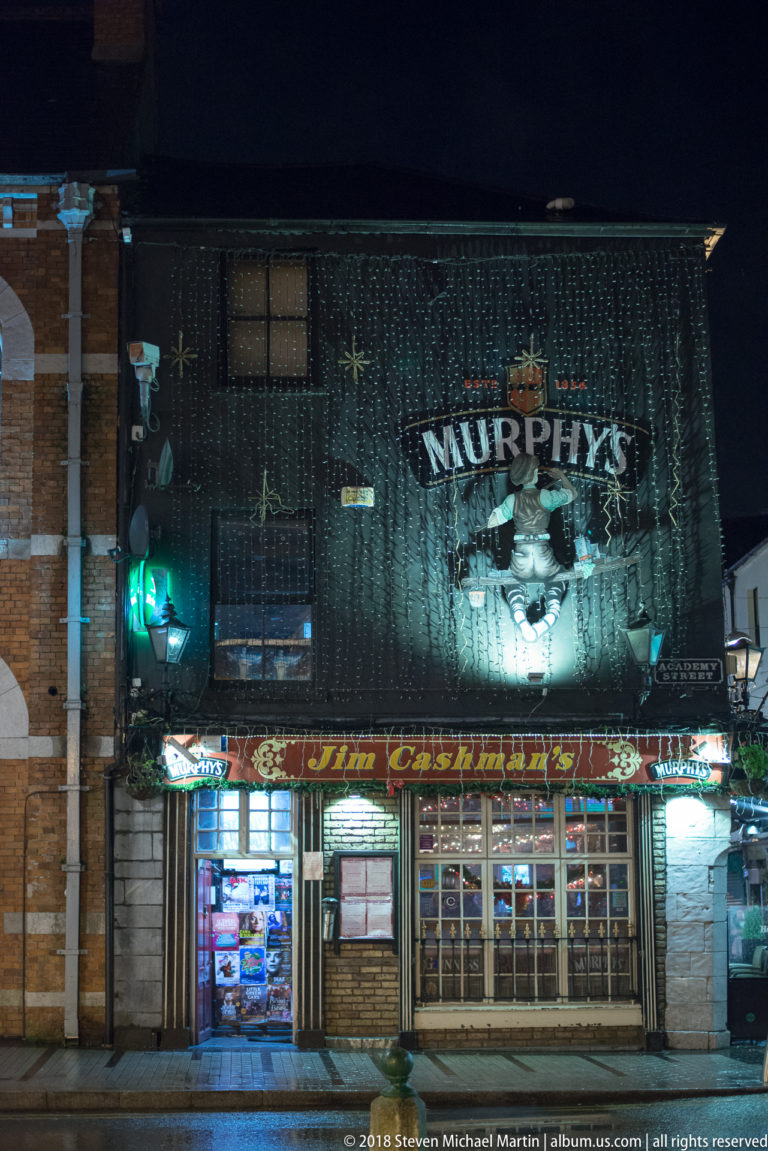 Murphy’s-branded pub façade with painted emblem, strings of lights, green lamps, and glazed frontage.