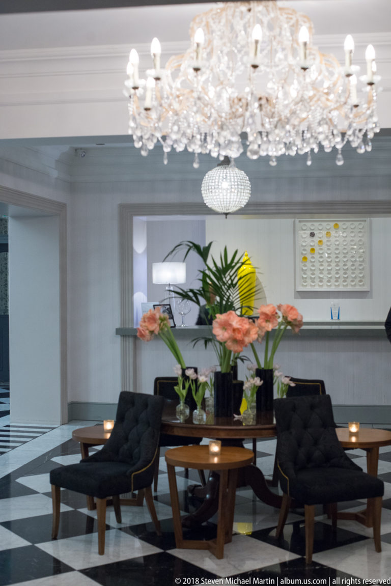 Crystal chandelier above black lounge chairs, wooden table, pink flowers, and black-and-white tiled floor.