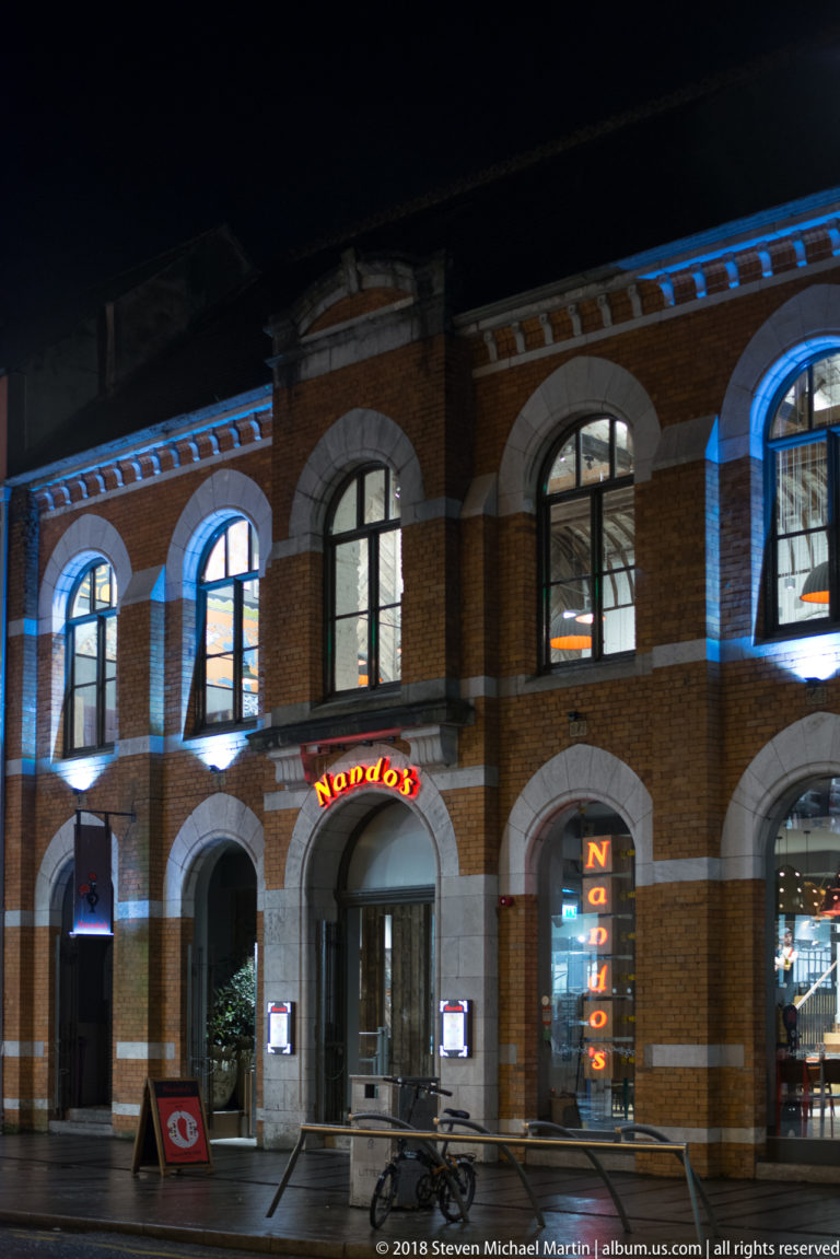 Nando’s entrance in a red-brick building with tall arched windows, pale stone trim, and blue night lighting.