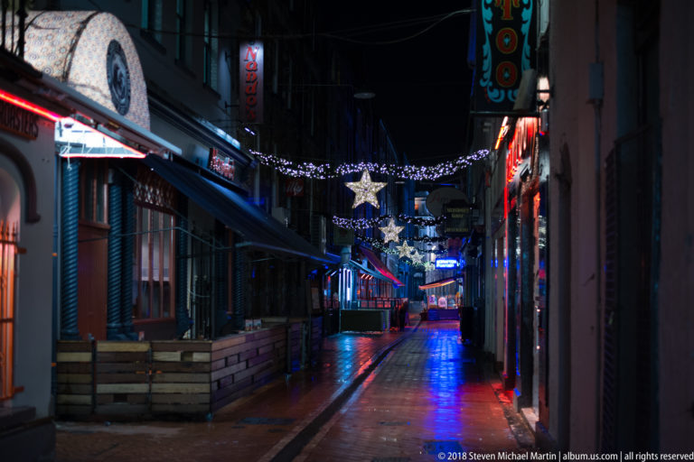 Narrow wet lane lined with bars, neon signs, outdoor seating, and hanging illuminated stars at night.