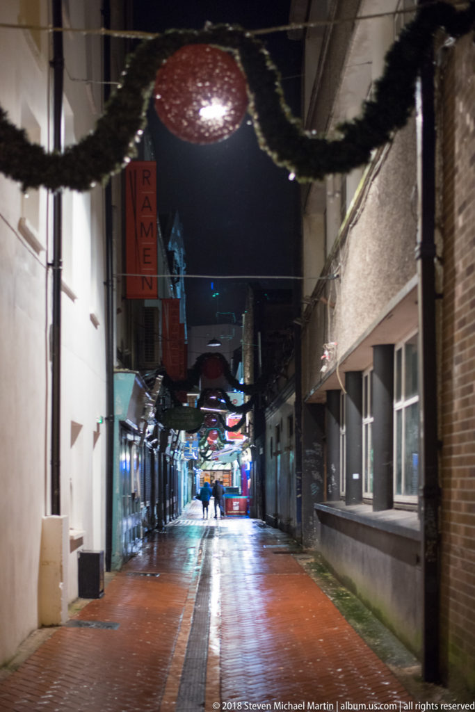Wet narrow lane with overhead garlands, glowing spherical decoration, shops, and distant pedestrians at night.