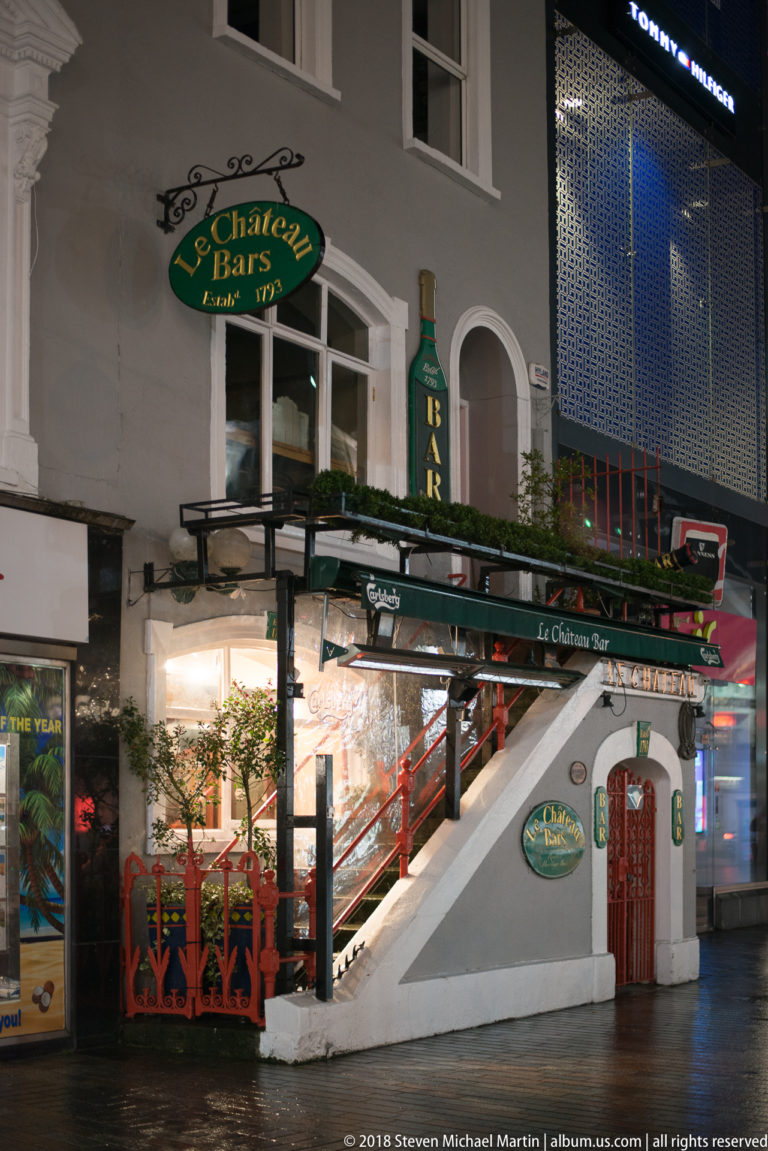 Le Chateau Bar with green signage, white façade, raised terrace, red railings, stairs, and planters at night.