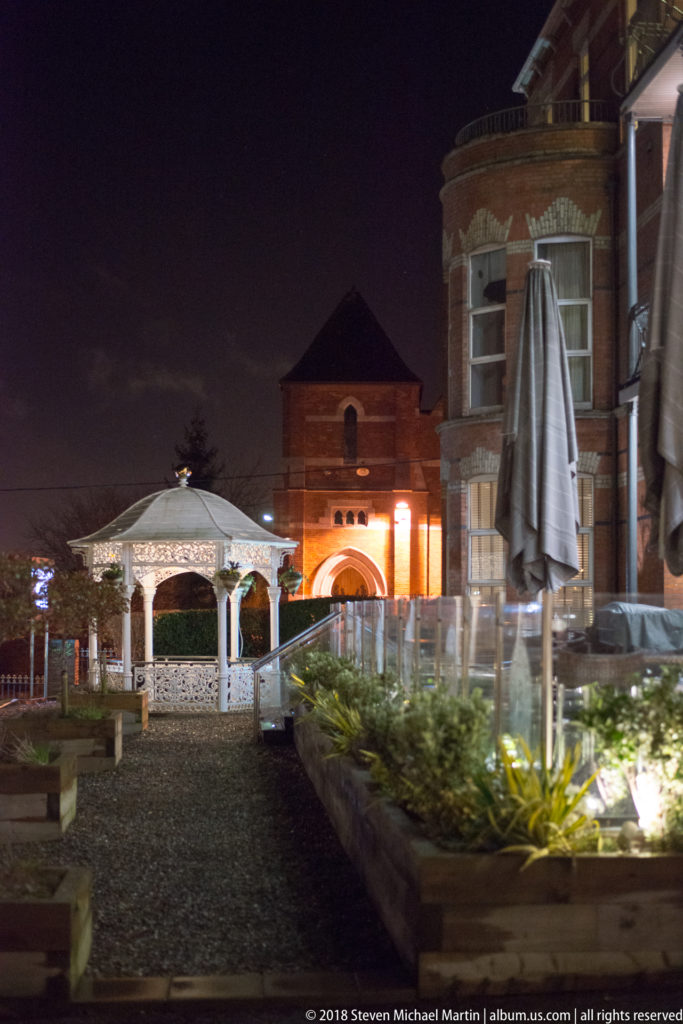 Nighttime brick courtyard with white gazebo, planted beds, paving, and warm exterior lighting.