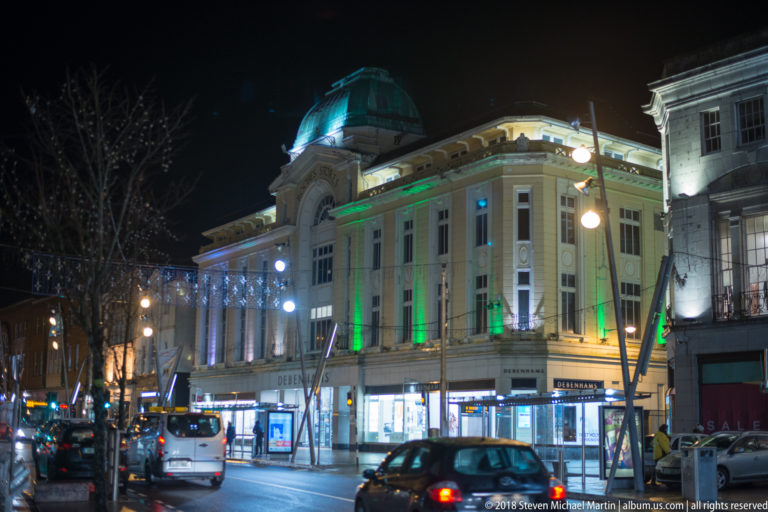 Large pale corner building with classical detailing, green lighting, domed roof feature, traffic, and shops at night.