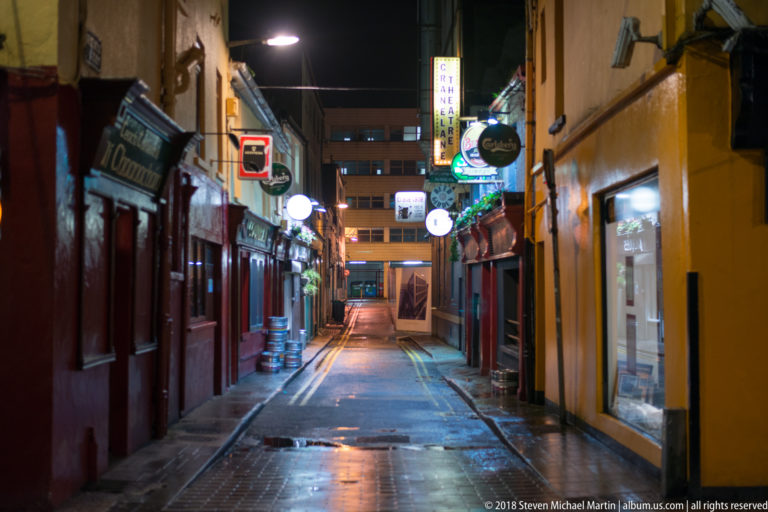 Narrow Cork street at night with wet pavement, pub signs, lit façades, beer kegs, and double yellow lines.