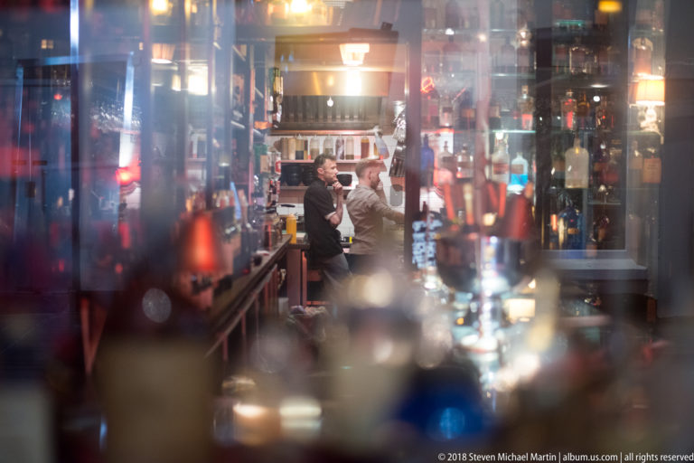 Two patrons talking at a Cork pub bar, framed through blurred bottles and reflective glass in warm interior light.