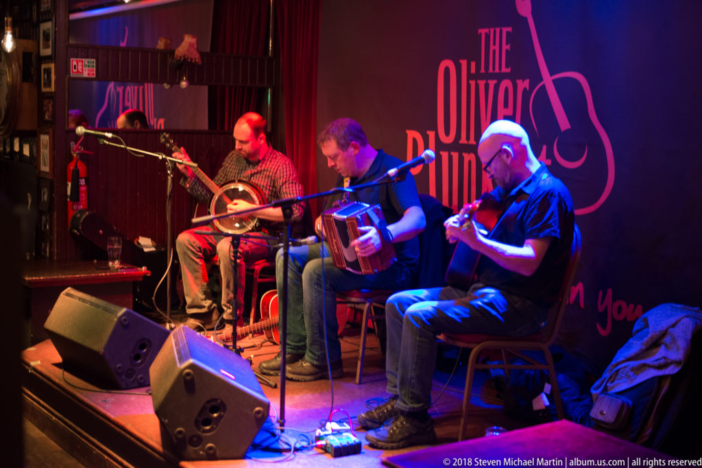 Four musicians seated onstage at The Oliver Plunkett in Cork, performing traditional music under red and blue lighting.