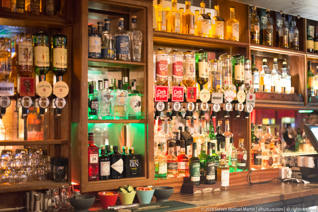 Illuminated Cork pub back bar filled with spirit bottles, draught taps, glassware, and bar tools in wood shelving.