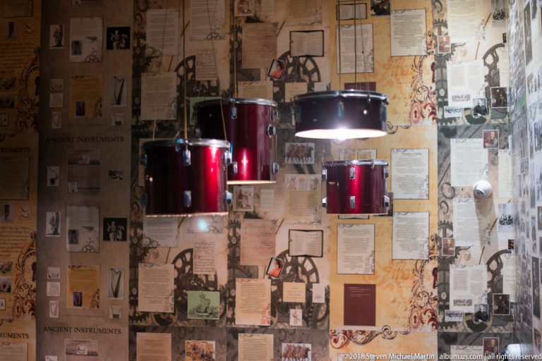 Red drum-shell pendant lights hanging before a wall covered with old prints, diagrams, advertisements, and documents.