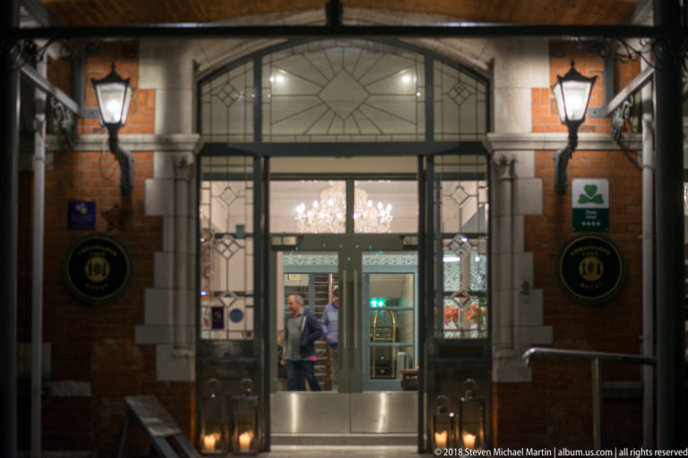 Glazed hotel entrance with pale stone surround, brick walls, lanterns, and illuminated interior beyond.