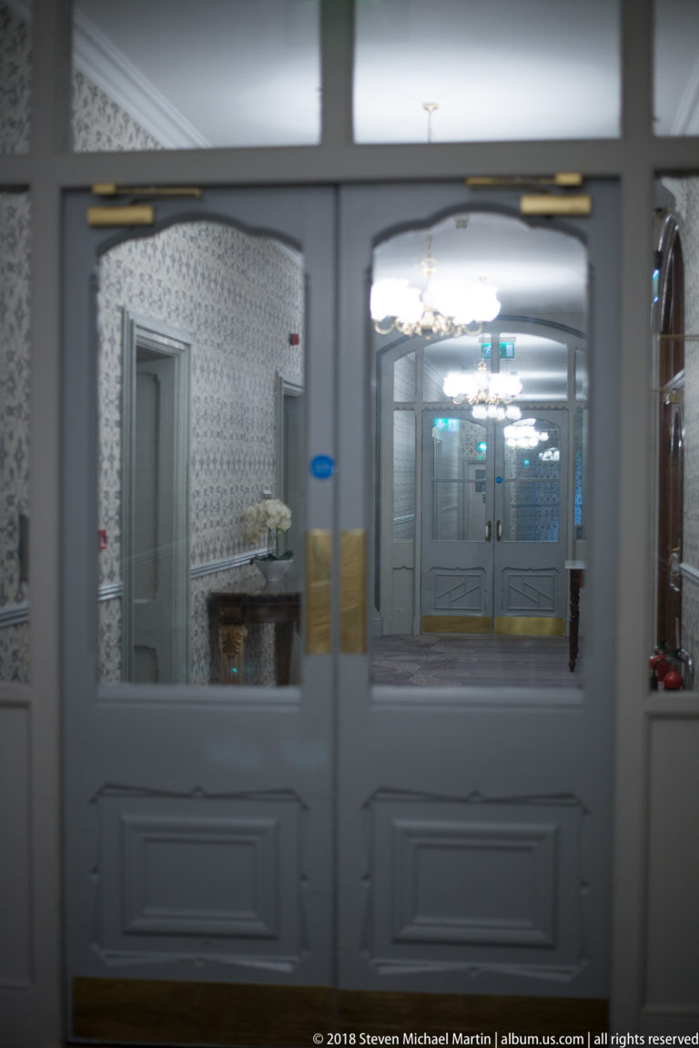 Glazed gray double doors with brass plates framing a bright interior of mirrors, patterned walls, and decorative lights.