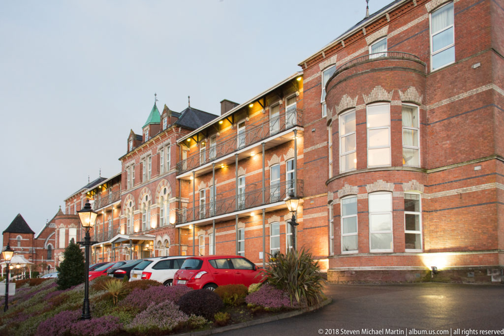Large illuminated red-brick building in Cork with projecting bays, balconies, stone trim, landscaped grounds, and parked cars.