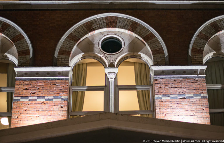 Paired round-arched windows with circular oculi, slender columns, and pale stone trim in a brick façade.