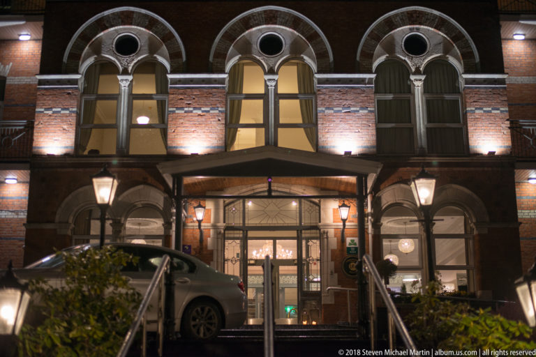 Illuminated red-brick hotel façade with arched windows, entrance canopy, steps, and parked car at night.