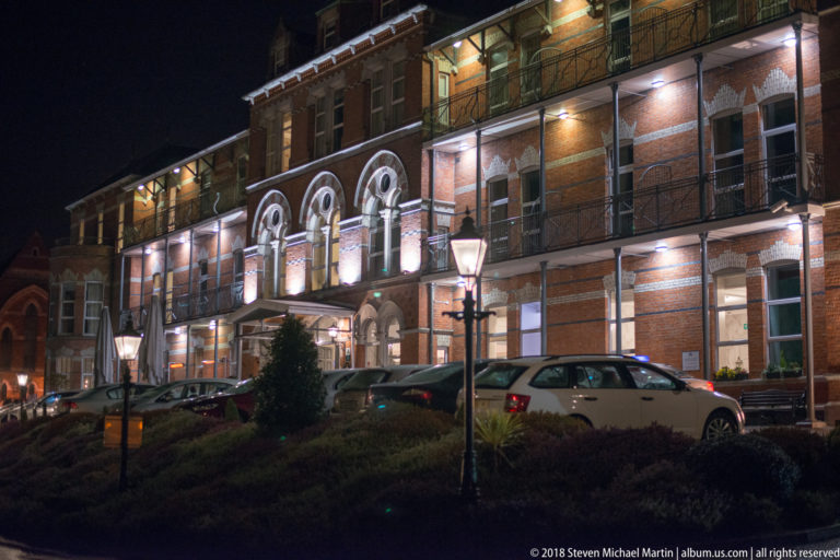 Long illuminated brick hotel façade with balconies, central arches, parked cars, and landscaped slope.